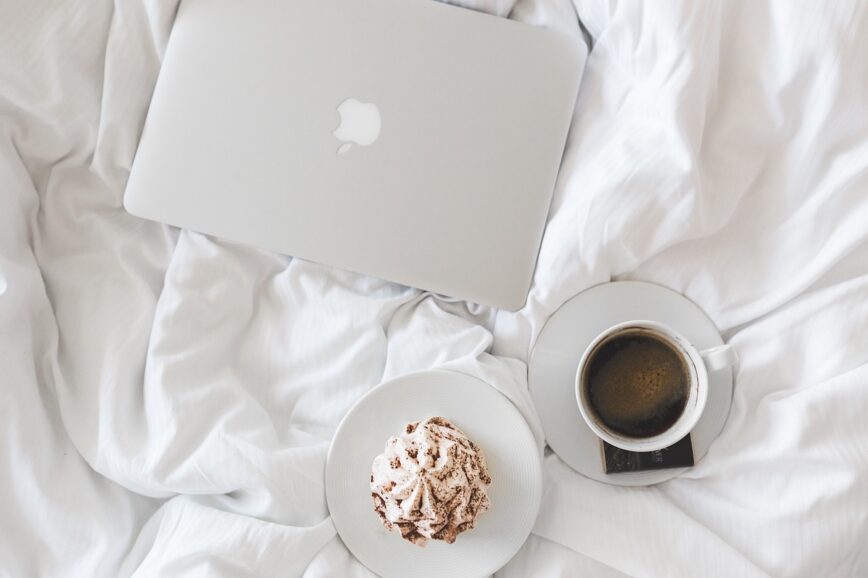 laptop on a bed with a coffee and baked good